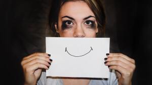 Woman holding a piece of paper with a laughing mouth over her sad face.